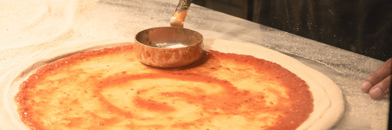 Image of someone putting sauce on pizza dough.