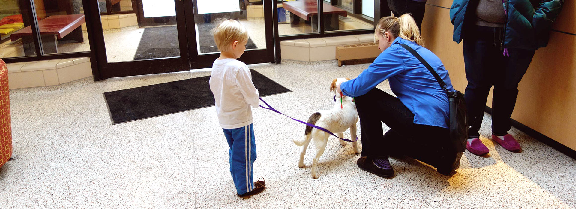A little boy standing with a dog on a leash. His mother is kneeling down petting the dog.