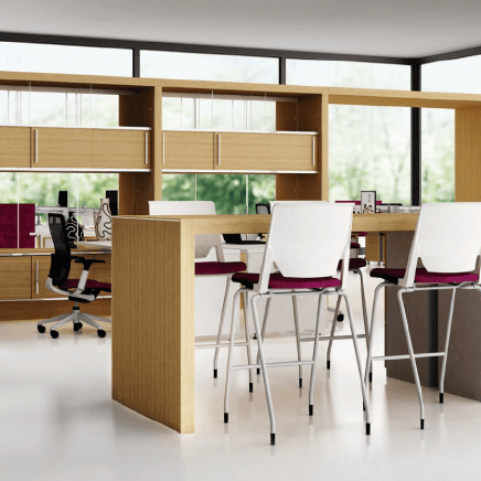 Open plan working space featuring a high-top working desk and stools, as well as desks, chairs and storage in front of windows in the background