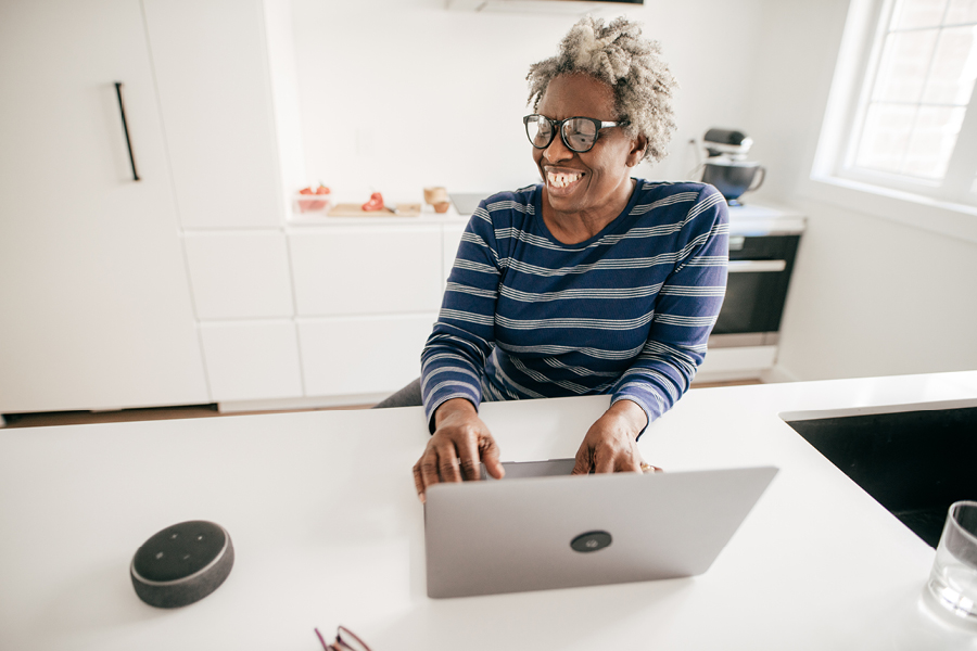A black woman working on her computer at her modern kitchen's countertop, gleefully looking over and providing voice commands to her Amazon Alexa device.